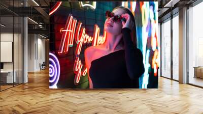 Cool young stylish woman model in fashion black clothes puts on sunglasses and walks near the colorful neon lights of the signs Wall mural