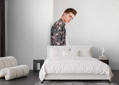 Elegant pretty young man with trendy hairstyle in white t-shirt in floral vintage shirt in stylish trousers is resting near white wall in studio. Attractive guy model in youth fashion clothes indoors. Wall mural