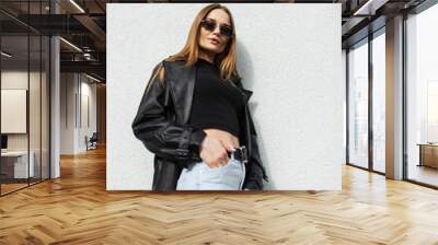 Fashionable beautiful young woman model with gold vintage sunglasses in a fashion long black leather coat with T-shirt and high-waisted jeans standing near the white wall on the street Wall mural