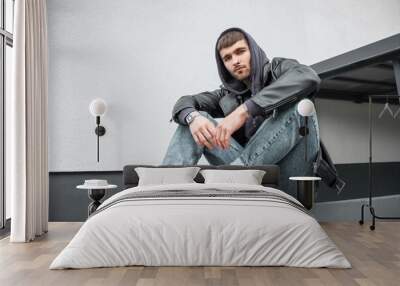 Fashionable handsome young hipster man wearing trendy clothes with jacket, hoodie, jeans and sneakers sitting near the white wall on the street Wall mural