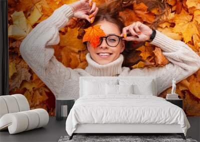 Funny young hipster woman with a positive smile with an orange maple leaf in glasses in a trendy knitted sweater lies on autumn foliage in the park. Joyful girl holds in a hand golden leaf near face. Wall mural