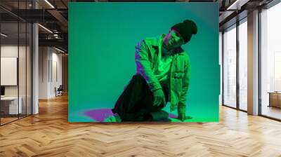 Handsome happy professional dancer man in fashionable denim clothes sits in the studio on a colored background with green and purple lights Wall mural