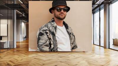 Handsome stylish hipster man with beard, sunglasses and bucket hat in fashion summer outwear with beach shirt near the wall on the street Wall mural