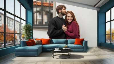 Happy beautiful young fashion couple in stylish casual clothes with hoodie standing and hugging in the city near a white wall. Handsome hipster man with a beard with a tattoo and a redhead girl smile Wall mural