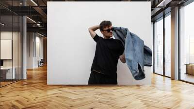 Modern young man hipster puts on a stylish denim jacket. Attractive guy in fashionable clothes in sunglasses posing on a street in the city near a white wall. Trendy new collection of men's clothing. Wall mural