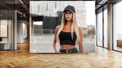 Pretty model hipster woman in a black baseball cap with a black stylish tank top and denim shorts with a belt stands in the city. Modern urban female style look outfit Wall mural