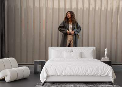 Pretty young stylish woman model with curly hair in a fashionable long coat with boots straightens her belt near a metal wall. Female casual fashion and style Wall mural