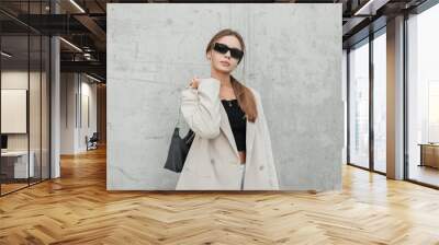 Stylish beautiful urban woman model with cool sunglasses in a fashionable beige blazer and a black top with a bag stands and poses near a gray concrete wall in the city Wall mural