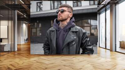 Stylish handsome young man hipster model in fashionable casual outfit with leather rock jacket and hoodie with sunglasses walking on the street Wall mural