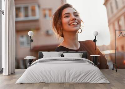 Young happy woman with a beautiful smile in a stylish T-shirt in a coat is standing and enjoying the orange sunset in the city near the houses. Joyful girl model relaxes in the sun. Sunny weekend. Wall mural