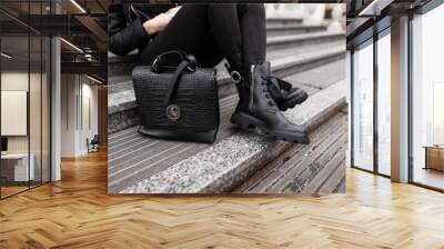 Young stylish woman in fashionable autumn black clothes sits on the steps in the city. Close-up of female legs in jeans in boots with a leather fashion handbag. Trendy women's shoes and accessories. Wall mural