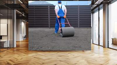 male worker in a uniform prepares a plot of land for sowing a lawn using a garden roller, leveling and compacting the earth Wall mural