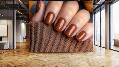 Close-up of a hand with beautifully manicured nails painted in a glossy brown shade, framed by a cozy brown knit sleeve. Autumn leaves and small pumpkins are in the background. Wall mural