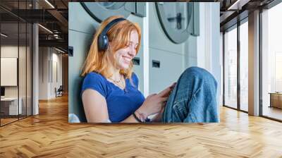 Smiling redhead girl listening to music in a public laundry waiting for her clothes to be washed Wall mural