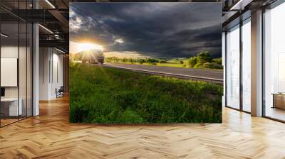 Blue truck driving on the asphalt road in rural landscape in the rays of the sunset with dark storm cloud Wall mural