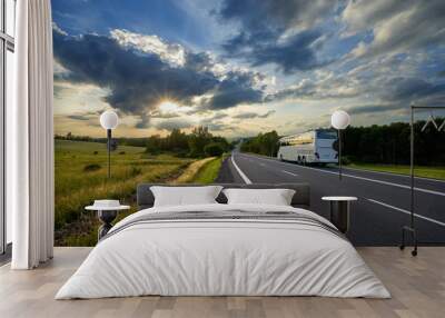 Bus traveling on the asphalt road in a rural landscape at sunset with dramatic clouds Wall mural