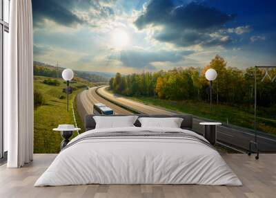 Three white buses traveling on the asphalt highway between deciduous forest in autumn colors under the radiant sun and dramatic clouds. View from above. Wall mural