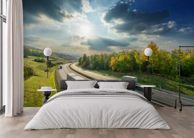 Trucks driving on the asphalt highway between deciduous forest in autumn colors under the radiant sun and dramatic clouds. View from above. Wall mural