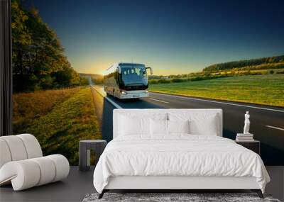 White bus traveling on the asphalt road around line of trees in rural landscape at sunset Wall mural