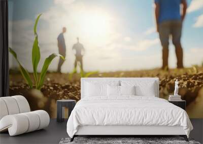 A close view of a small green plant growing in dry soil, with two figures discussing in the background under a bright, sunny sky. Wall mural