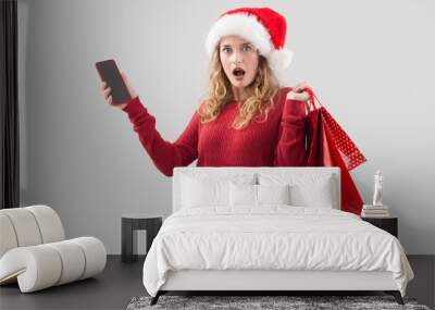 Amazed merry Christmas woman wearing Santa hat holds cellphone and red shopping bags. Image for sale offer promotions, online shopping, advertising banner, poster, or gift card, isolated on background Wall mural