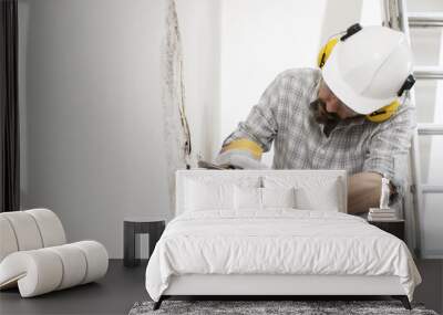 house renovation concept, construction worker breaks the old plaster of the wall with pneumatic air hammer chisel, wears gloves, helmet and safety yellow headphones , close up with white copy space Wall mural