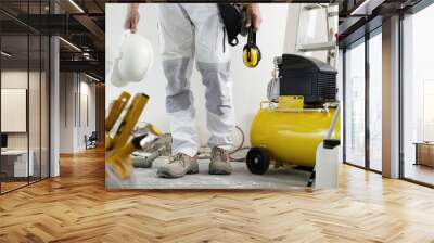 house renovation concept, construction worker holds in hands helmet and safety headphones, air compressor and construction tools in the background Wall mural