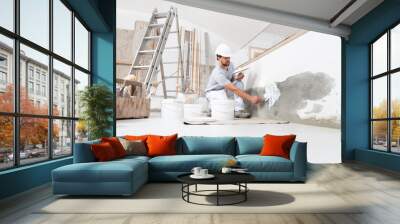 man plasterer construction worker at work, takes plaster from bucket and puts it on trowel to plaster the wall, wears helmet inside the building site of a house Wall mural