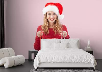 Smiling blonde young woman opening a Christmas present shopping bag while wearing a Santa hat, isolated on a pink background. For sale and offer promotions, online shopping adv banner, or gift card Wall mural