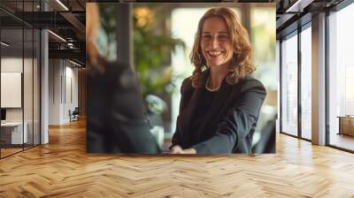 A happy mid-aged businesswoman manager smiling and shaking hands with a client in her office, celebrating a successful deal while standing at the meeting table. Wall mural