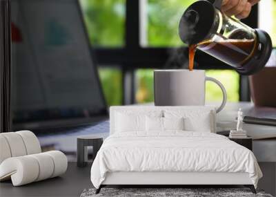 A hand pouring steaming coffee in to a cup on a work desk when work from home Wall mural