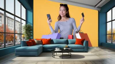 Beautiful young Asian woman with colourful shopping bags with smart phone and credit card isolated on yellow background Wall mural