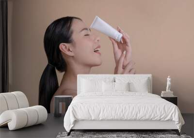 Portrait of a young Asian woman smiling holding mockup product for advertising text place, beige background. Concept of healthcare for skin, beauty care product for advertising. Wall mural