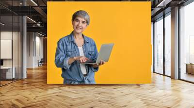 Portrait of middle aged Asian woman 50s wearing casual denim shirt white t-shirt holding laptop computer and pointing fingers isolated on colour background, looking and smiling at camera Wall mural