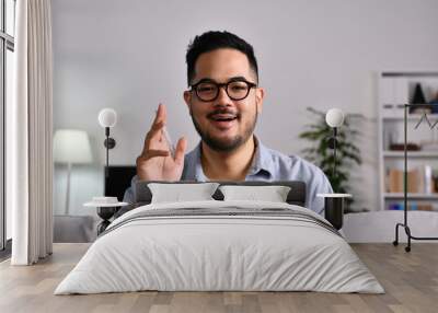 Portrait shot of happy friendly and confident Asian businessman sitting on a couch at home, looking and talking at camera and waving hand saying hello during video call Wall mural