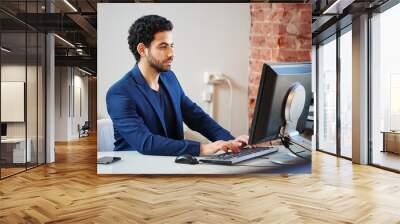 An Arab man in a jacket sitting in the workplace at the computer and typing on keyboard in office Wall mural