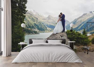 Beautiful wedding photo on mountain lake. Happy Asian couple in love, bride in white dress and groom in suit are photographed against background of the Kazakh landscape Wall mural