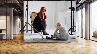 Girl photographer photographing fashion model in black sitting on a chair on white background in Studio Wall mural