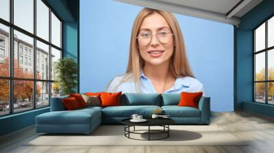 portrait beautiful young blond woman in business shirt with glasses toothy smiling and looking at the camera isolated on blue studio background with copy space Wall mural