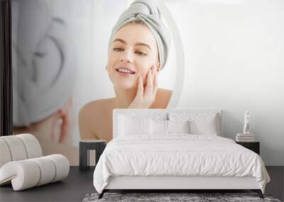 Portrait of  young girl with  towel on head in white bathroom looks and touches her face in the mirror and enjoys youth and hydration. Natural beauty, home care for problem skin Wall mural