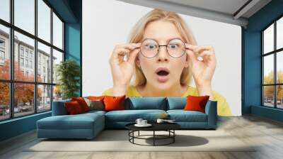 Surprised woman in round glasses with open mouth and bulging eyes looks into camera and sees something incredible and amazing isolated on white background Wall mural