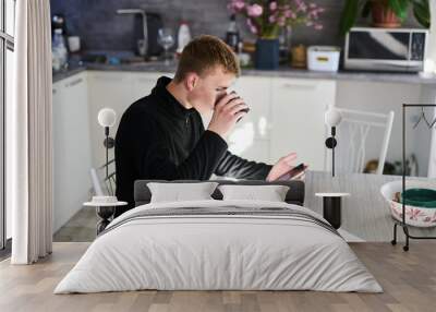 Young man sitting in the kitchen in the morning, drinking coffee and checking the news on his smartphone Wall mural