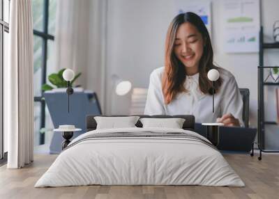 Asian businesswoman is smiling while working on documents at her desk in her bright modern office Wall mural