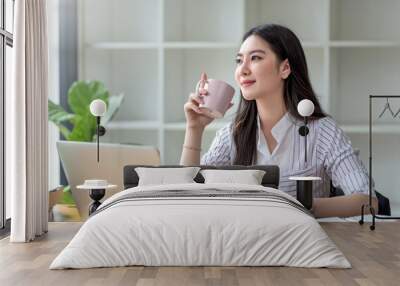 Beautiful young Asian businesswoman smiling holding a coffee mug and laptop working at the office. Wall mural