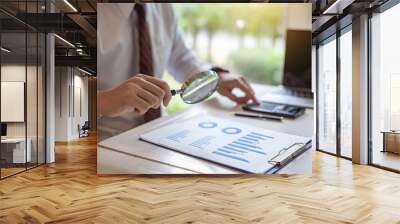 Businessman holding a magnifying glass with graph information document on the desk. Wall mural