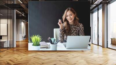 Charming asian businesswoman sitting working on laptop in office. Looking at camera. Wall mural