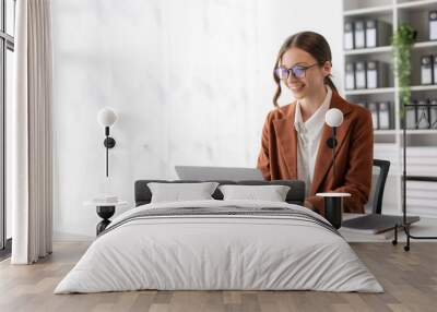 Charming businesswoman sitting working on laptop computer in office. Smiling female employee executive typing message using laptop computer for business in modern office. Wall mural