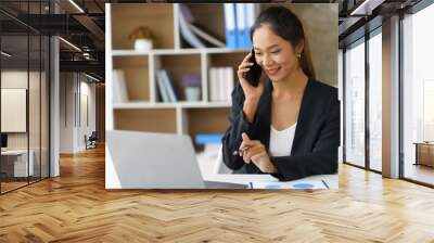 Happy young Asian business woman sitting smiling and talking on mobile phone in office. Wall mural