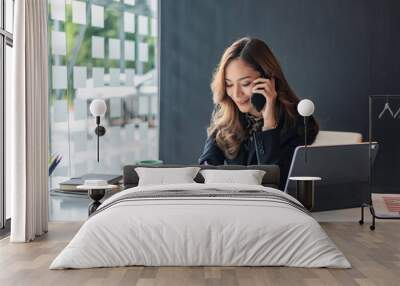 Happy young asian woman talking on the mobile phone and smiling while sitting at her working place in office. Wall mural