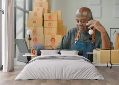 Man wearing apron at desk with tablet, on phone, writing in notebook. Stack of parcels behind him, engaged in small business activities. Wall mural